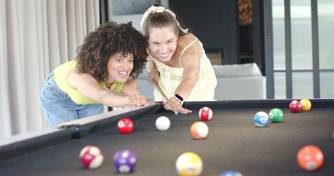 Diverse women playing pool at home laughing while lining up cue shot on black felt table