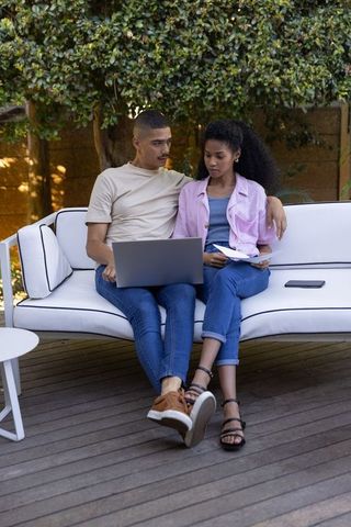 Young couple reviewing finances on laptop while sitting on patio sofa