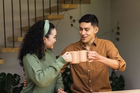 Young couple clinking beige mugs on wooden counter sharing cozy morning coffee toast