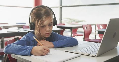Focused Young Girl Writing Notes with Laptop and Headphones