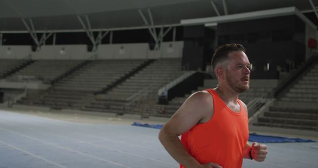 Man running on indoor track in stadium wearing orange tank top, embodying focus and determination. Useful for sports promotions, fitness blogs, and health campaigns.