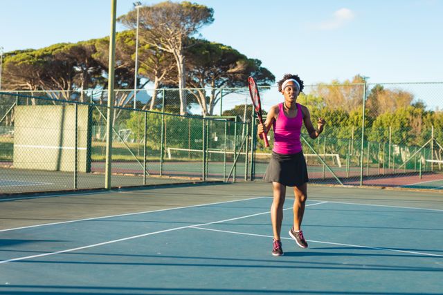 Full length of excited young female african american tennis cheering herself at court on sunny day. unaltered, sport, competition and tennis game concept.