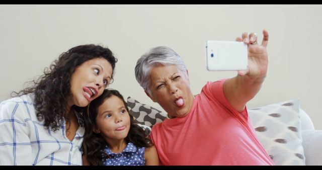 Mother, daughter, and grandmother sitting on couch, making silly faces while taking selfie with smartphone. This captures a light-hearted and joyful moment, perfect for advertisements, family-centric social media content, or articles on family relationships, bonding, and multicultural families.