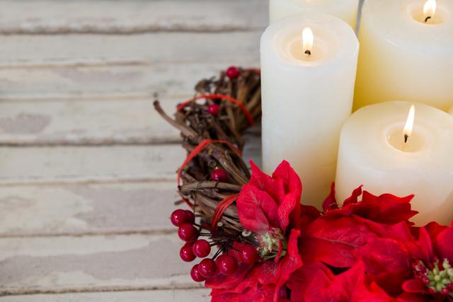 This image shows a festive arrangement of white candles surrounded by a wreath made of poinsettia flowers and red berries on a wooden plank. Ideal for use in holiday greeting cards, Christmas-themed advertisements, seasonal blog posts, or social media content celebrating the winter holidays.
