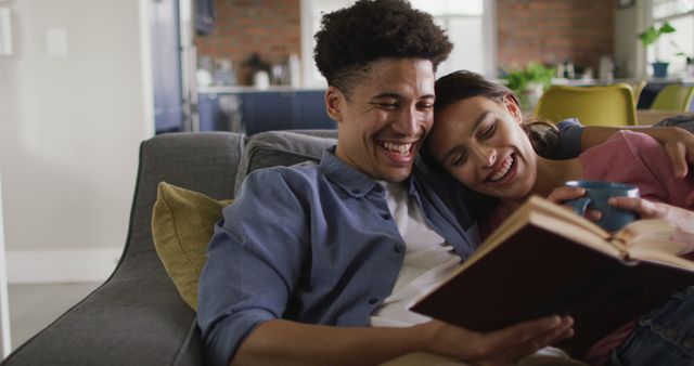 Happy biracial couple sitting on sofa in living room, reading book and drinking coffee. quality time, relaxing together at home.