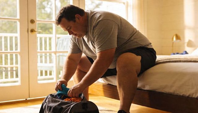Man packing duffel bag on bed in sunlit bedroom preparing weekend trip