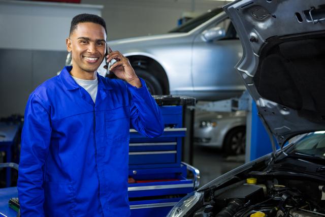 Mechanic in blue uniform smiling while talking on mobile phone in repair garage. Ideal for use in automotive service advertisements, customer service promotions, and technology in the workplace content.