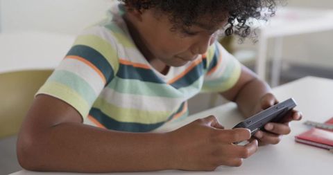 African american boy holding handheld game console at classroom desk, focusing on screen