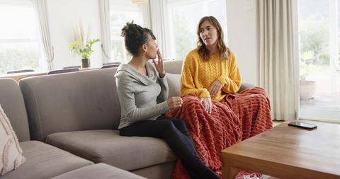 Mid-adult women chatting on sofa wrapped in cozy throw during relaxed living room talk