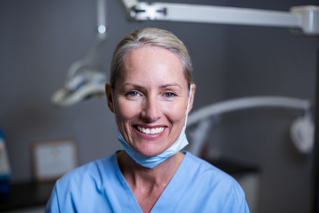 Female dental assistant smiling in a clinic, wearing light blue scrubs and a protective mask around her neck. Suitable for use in healthcare advertising, dental care promotions, medical staff recruitment, and professional healthcare websites.