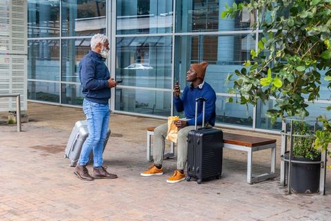African american travelers chatting and waiting with carry-on luggage on urban plaza bench using pho
