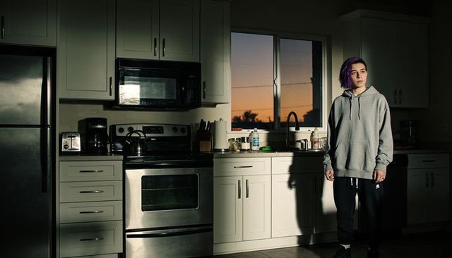 Young person with purple hair standing in kitchen at sunset wearing hoodie casting shadow