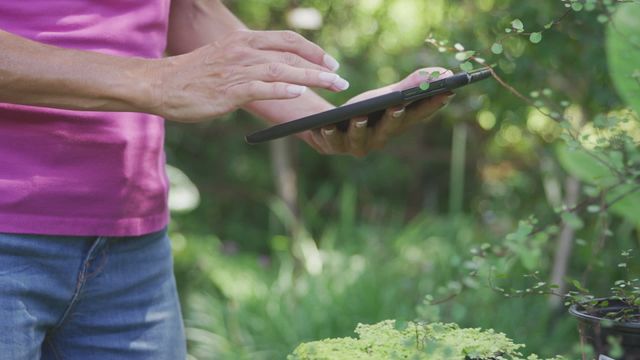 This portrayal of a woman in a garden demonstrates modern technology use in an outdoor setting. The tablet suggests possible interactions with gardening apps or online resources for plant care, suitable for illustrating eco-friendly lifestyles, mobile technology in learning, and women's involvement in gardening.