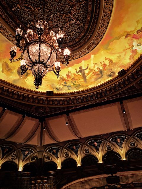 Intricately decorated theater ceiling features a grand chandelier, beautifully painted fresco, and elaborate molding. Ideal for use in articles or websites focusing on historical architecture, theaters, cultural spaces, or luxury interior designs. Also suitable for travel guides or blogs highlighting must-see historic buildings.