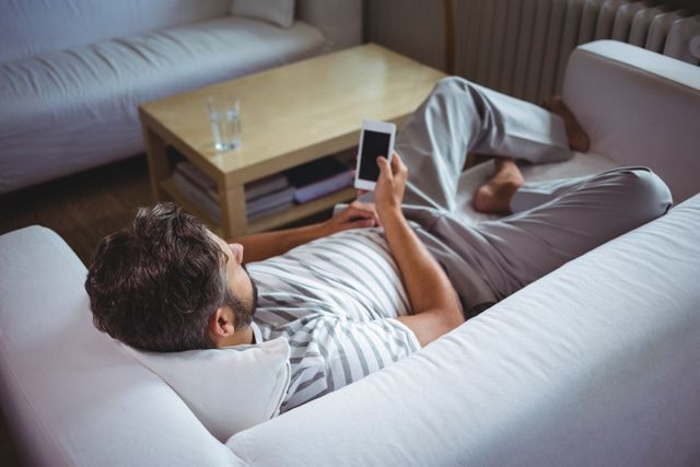 Man lying on a white couch in a living room, using a smartphone. Ideal for themes related to home comfort, technology use, relaxation, and modern lifestyle. Suitable for articles, blogs, and advertisements focusing on home living, tech gadgets, and personal leisure time.