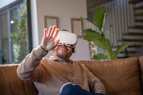 Mature asian man using virtual reality headset on leather sofa smiling and reaching