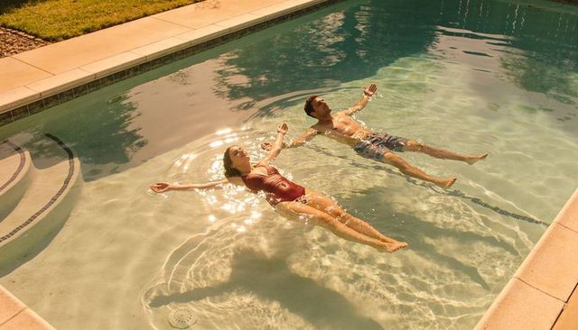 Couple floating in sunlit backyard pool relaxing near steps and shallow water