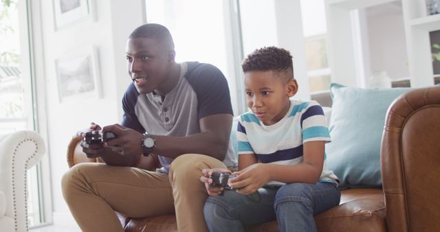 Image of happy african american father and son sitting on sofa and playing image games. Family, spending quality time together at home with technology concept.