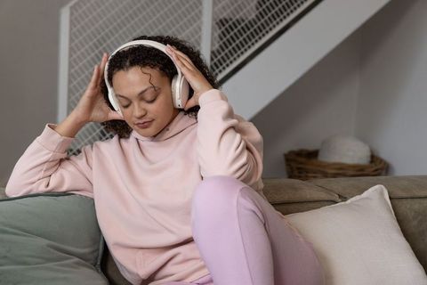 Woman listening to music on white headphones relaxing on pastel sofa in cozy home lounge