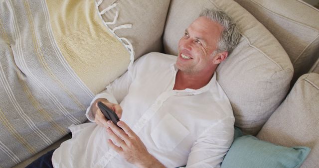 Happy caucasian man lying on sofa in living room, using smartphone. Spending time at home alone.