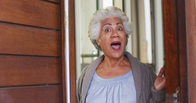 Senior woman with grey hair standing in doorway looking surprised. Wearing a cardigan and light blue top. Suitable for use in articles about aging, senior living, and expressions of surprise.