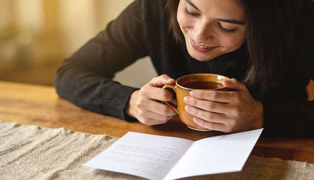 Smiling woman cradling warm ceramic mug while reading handwritten letter at wooden table