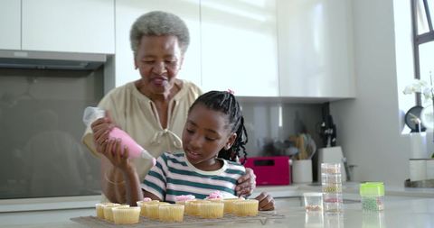 Grandmother guiding granddaughter decorating cupcakes with pink frosting in modern kitchen