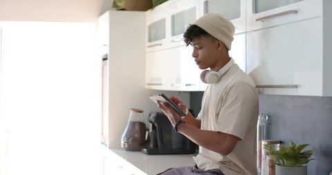Young man using tablet in sunlit modern kitchen with headphones, coffee maker and plant