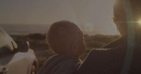 Mother holding baby at coastal road by car with sunset flare