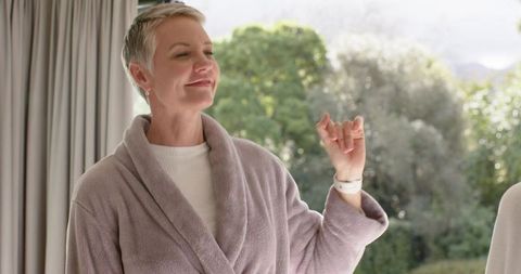 Senior woman in purple bathrobe raising hand by glass door enjoying serene morning at home