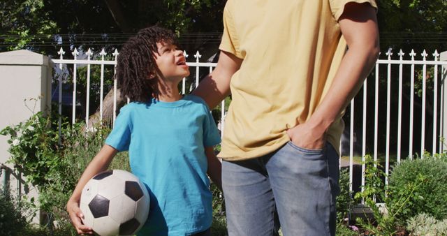 Father wearing casual clothes bonding with son outdoors while holding a soccer ball. Perfect for themes emphasizing family connections, parent-child activities, outdoor play, and childhood joy.