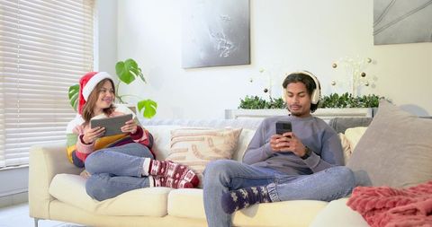 Couple Relaxing on Sofa Wearing Santa Hat and Headphones Using Tablet and Smartphone