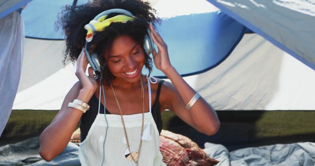 Young woman with curly hair smiling while listening to music on headphones inside a tent. Perfect for themes related to summer, outdoor adventures, camping, relaxation, or a carefree lifestyle. Useful for advertisements, lifestyle blogs, or travel-related content.