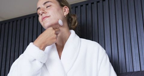 Female in white robe applying rose quartz facial roller to jawline for calm and self care