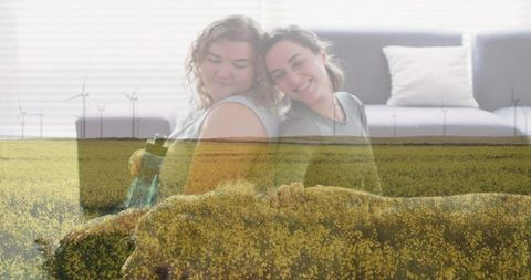 Two women sharing relaxed back-to-back moment over canola field and wind turbines overlay