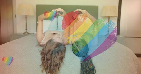 Two women lying on bed sharing smartphones with rainbow heart overlay, lgbtq pride