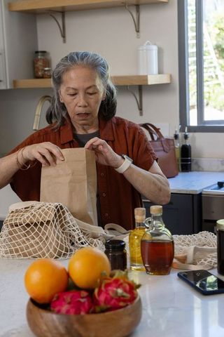 Senior asian woman packing groceries at kitchen counter with fresh produce and reusable bags