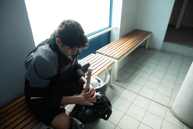 Caucasian teenage male field hockey player sitting on a bench in a locker room, focusing and preparing before a game. Ideal for use in sports-related articles, advertisements for athletic gear, or motivational content about focus and preparation in sports.