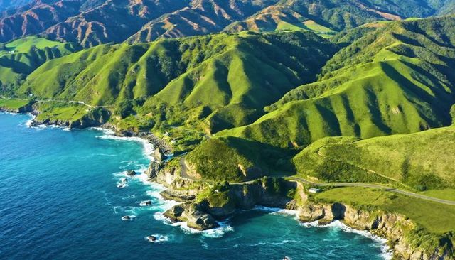 Aerial winding coastal road carving green headlands above turquoise ocean waves