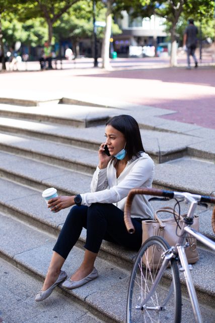 African American woman sitting on outdoor stairs, talking on smartphone, holding coffee cup, with face mask lowered. Bicycle and handbag beside her. Suitable for depicting urban lifestyle, pandemic-related themes, remote work, and modern living.