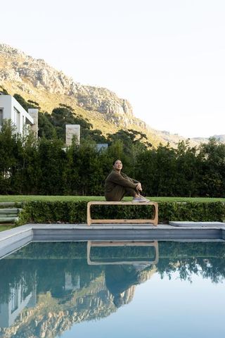 Mid adult asian woman sitting on bench by pool wearing olive-green outfit with mountain reflection
