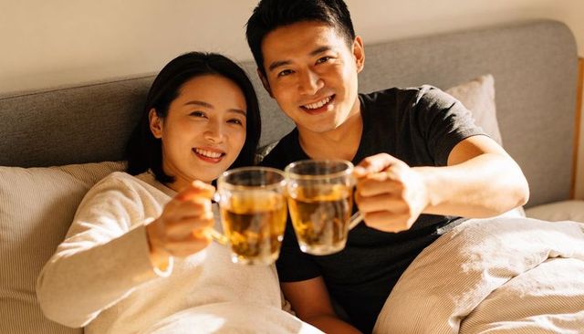 Cozy couple clinking clear glass mugs while reclining in bed under warm golden light