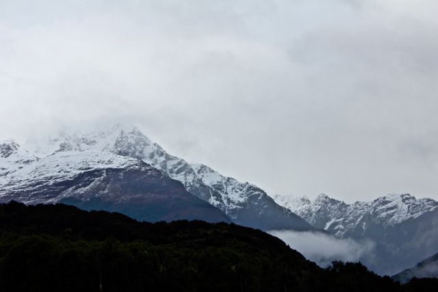 This image showcases snow-covered mountain peaks set against a cloudy sky, providing a serene and picturesque scene. The dark, dense foliage at the base of the mountains contrasts beautifully with the white snow, making it a stunning example of nature's beauty. This image can be used for nature-themed content, travel brochures, winter sports promotions, or as a desktop wallpaper.