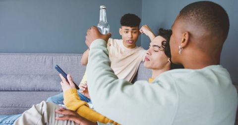 Multicultural young men lounging on gray sofa holding bottle and smartphone