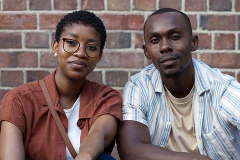 African couple sitting on brick wall, leaning toward camera, casual urban daytime portrait