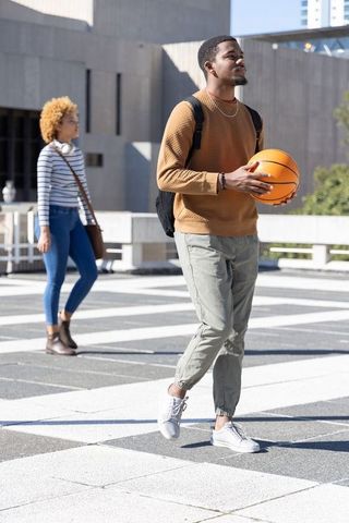 African american man holding basketball walking through urban campus plaza with student