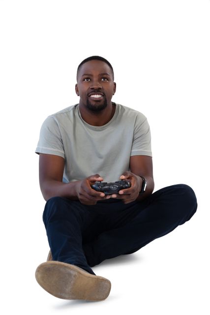 Young African American man sitting on floor, smiling while playing video game with controller. Ideal for use in advertisements, articles, and blogs related to gaming, technology, leisure activities, and young adult lifestyles.