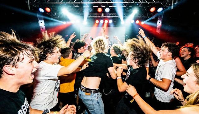 Blond fan jumping and headbanging in intense mosh pit under colorful stage lights