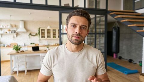 Man speaking to camera in modern open-plan loft kitchen with home gym, laptop workspace