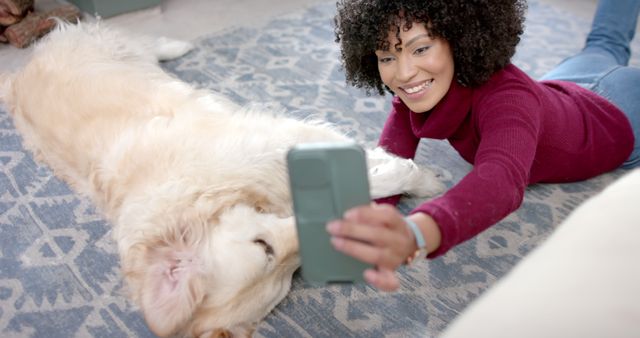 Woman lies down next to Golden Retriever on patterned carpet, taking selfie with smartphone. Suitable for themes of pet ownership, indoor lifestyle, joy and bonding between pets and owners. Great for use in social media posts, pet-related advertisements, and articles on home life.